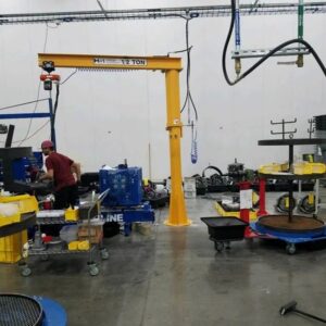 Yellow foundationless freestanding jib crane with electric hoist positioned in a production area with work tables and industrial equipment.