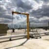 Outdoor foundationless freestanding jib crane with chain hoist installed on an industrial rooftop among HVAC units and piping.